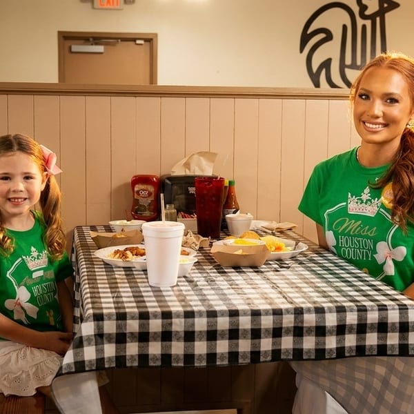 Two smiling females, a woman and a young girl, wearing green shirts, seated at a checkered table with food.