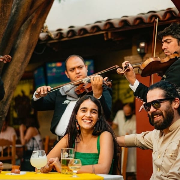 Mariachi band playing violins for smiling guests enjoying drinks at an outdoor restaurant.