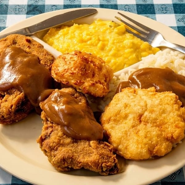 A plate of fried chicken with gravy, rice, corn, and biscuits on a blue and white checkered tablecloth.
