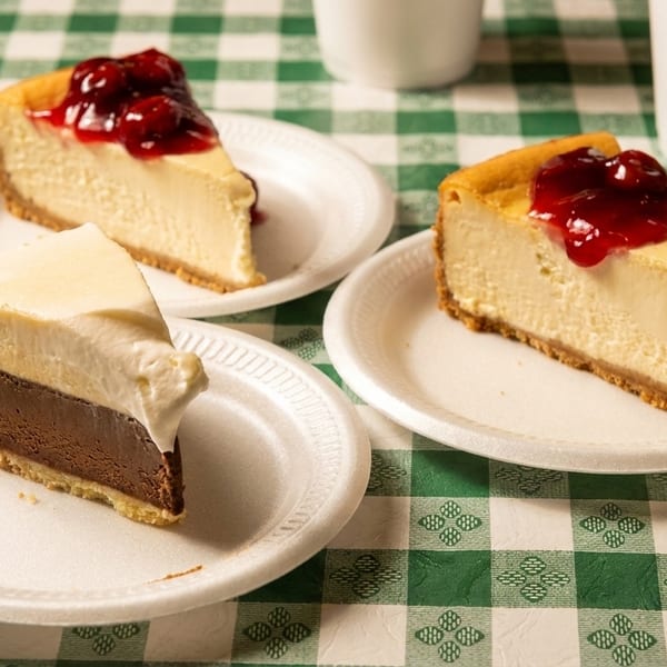 Three slices of cheesecake, two with cherry topping and one chocolate, on a green checkered tablecloth.