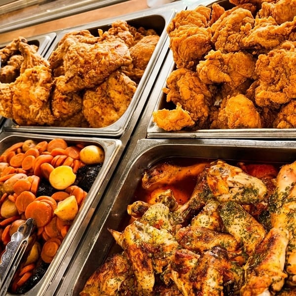 Buffet line with trays of fried chicken, roasted chicken, rice, and cooked carrots and potatoes.